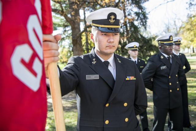A cadet stands at attention in full uniform with a scarlet and gray Columbus flag.