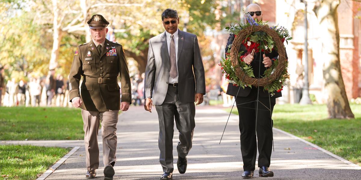 Three individuals walking towards the camera, one with a wreath in hand