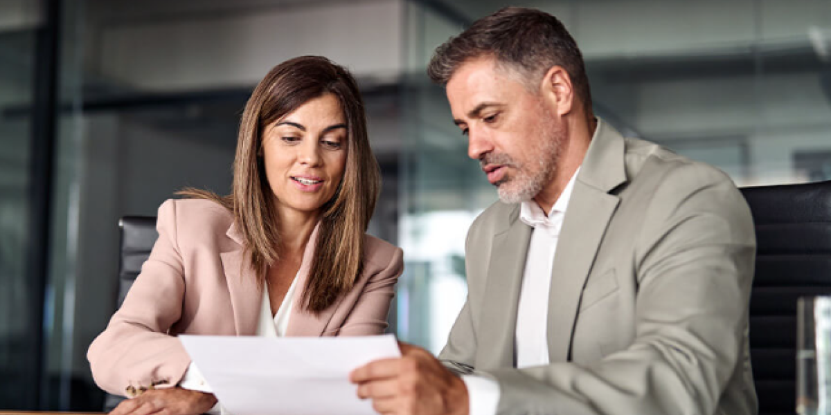 Two professionals, a man and a woman, look over paperwork while seated in a medium sized conference room.