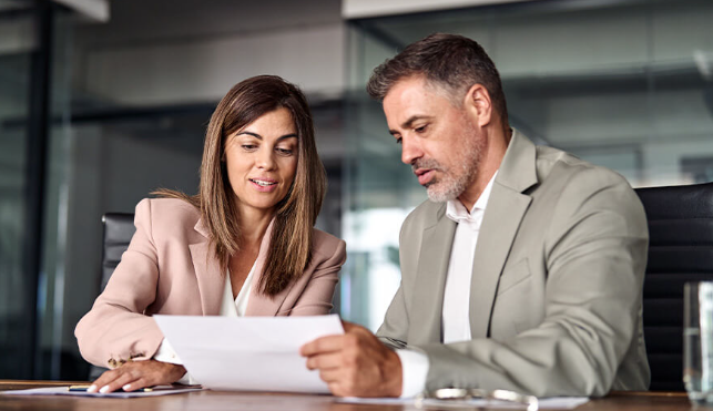 Two professionals, a man and a woman, look over paperwork while seated in a medium sized conference room.